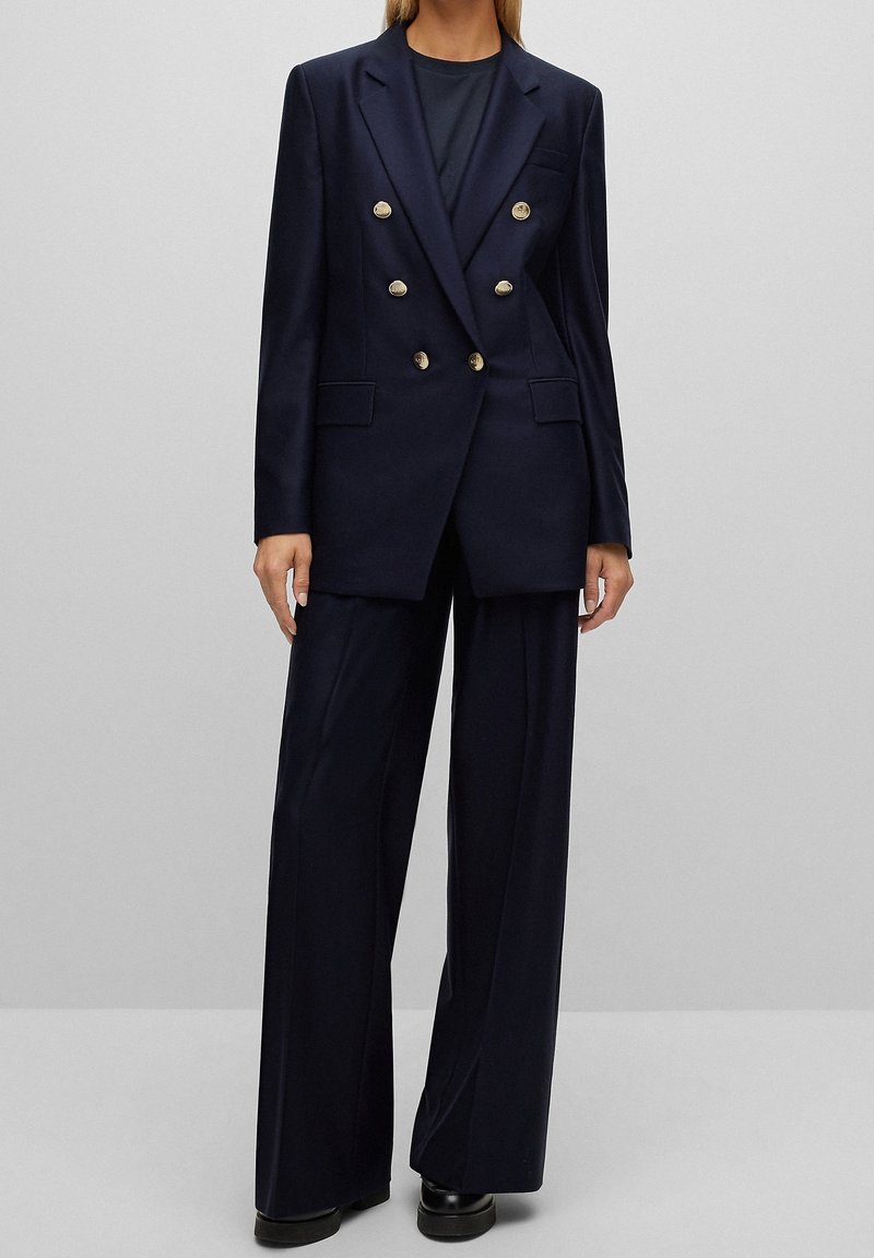 Veste bleu marine ajustée avec des boutons dorés croisés, revers à crans et ourlet pointu, associée à un pantalon large bleu marine. Texture lisse.