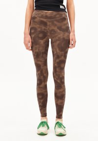 Een model staat in bruine, gemusterde leggings en witte sneakers met groene accenten, gedragen met een zwarte cropped top.