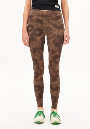 Una modella indossa leggings marroni con un motivo e sneakers bianche con accenti verdi, abbinati a un top nero corto.