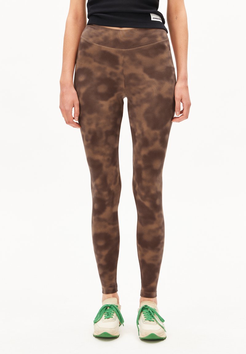 Een model staat in bruine, gemusterde leggings en witte sneakers met groene accenten, gedragen met een zwarte cropped top.