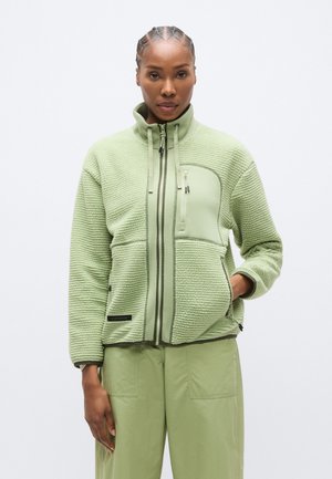 Zelená fleecová bunda s vysokým golierom, štruktúrovanou tkaninou, dvoma prednými vreckami so zipsom a kontrastným panelom na hrudi.