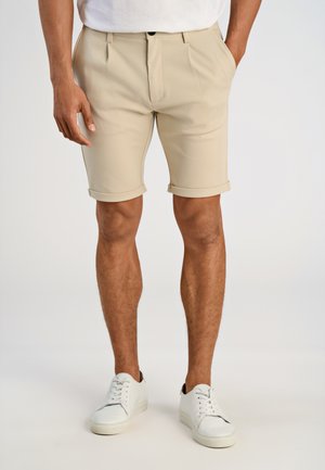 Beige tailored shorts with a front pleat, cuffed hem, and side pockets, paired with white sneakers and a plain white t-shirt.
