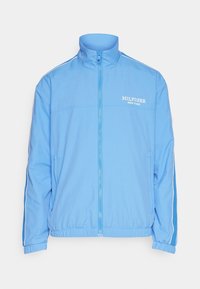 Chaqueta azul claro con cremallera hecha de tela lisa. Cuenta con cuello alto, puños elásticos, logo blanco en el pecho y bolsillos laterales.