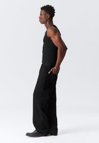 Svart tanktop og svarte vide jeans, kombinert med mørke støvler. Modellen står sidelangs og viser en tettsittende overdel og en løs underdel.