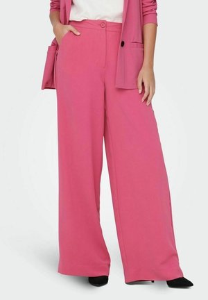 Femme portant un pantalon rose taille haute à jambes larges, associé à un blazer rose assorti et des chaussures noires à bouts pointus.