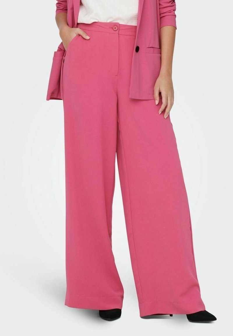 Femme portant un pantalon rose taille haute à jambes larges, associé à un blazer rose assorti et des chaussures noires à bouts pointus.