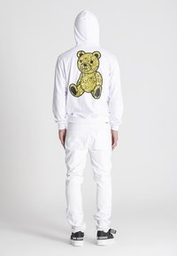 Biela mikina s veľkým žltým grafikom medveďa na chrbte, v kombinácii s bielymi nohavicami a čiernymi teniskami s logom.
