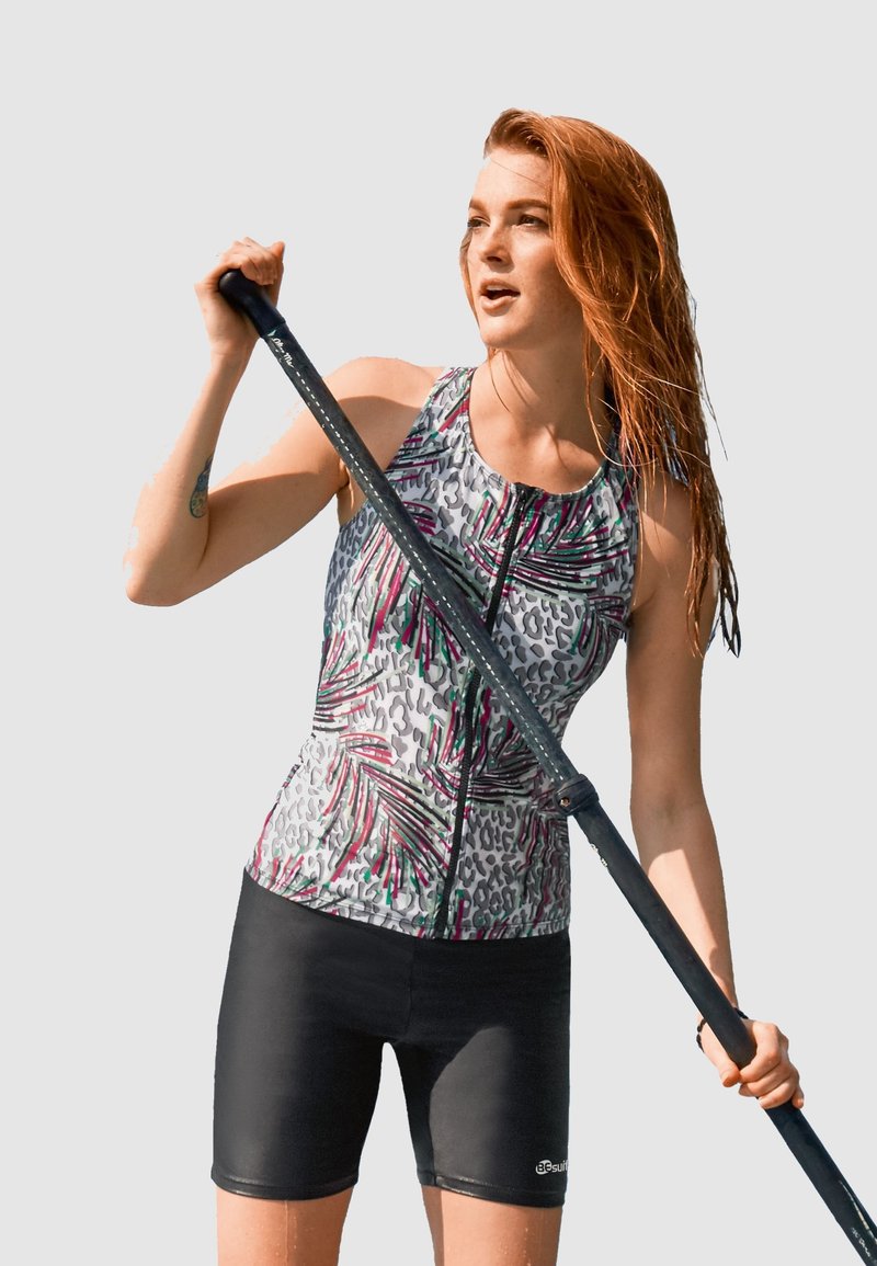 Maillot de bain en tissu coloré et à motifs, avec des motifs de léopard et de palmiers ; comporte une fermeture éclair à l'avant et un short noir. Une pagaie est tenue dans une main.