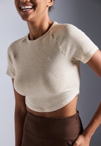 Bežasti rebrasti crop top s kratkim rukavima i okruglim izrezom, uparen s smeškastim uskim donjim dijelom s visokim strukom. Suptilni detalj loga na prsima.