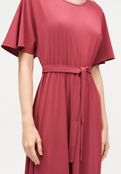 Γυναίκα που φοράει κοντομάνικο, ρούχο σε στυλ φορέματος, σε σκότεινο ροζ χρώμα με ζώνη και στρογγυλή λαιμόκοψη, μπροστά σε απλό ανοιχτό φόντο.