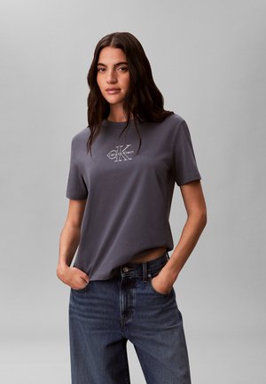 Femme aux longs cheveux foncés portant un t-shirt gris Calvin Klein et un jean bleu, debout les mains dans les poches devant un fond uni.
