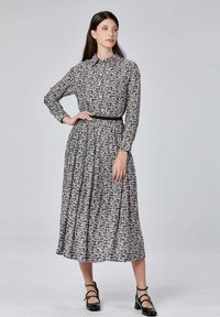 Femme debout, les mains sur les hanches, portant une robe midi noire et blanche à fleurs à manches longues et des chaussures à bride de cheville noires sur un fond gris uni.