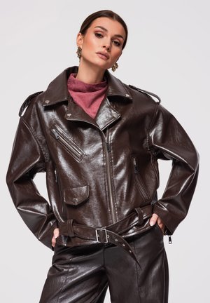 Femme portant une veste en cuir brun foncé brillante et un pantalon, associée à un haut bordeaux, posant les mains dans les poches devant un fond uni.