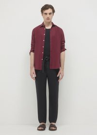 Chemise bordeaux boutonnée portée sur un t-shirt noir, associée à un pantalon noir et des sandales marron, avec un détail de taille nouée.