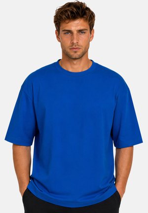Man met warrig bruin haar, draagt een los vallend koningsblauw T-shirt en zwarte broek, staat met handen in de zakken tegen een effen achtergrond.