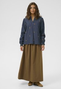 A farmerblúz bronz színű pillangó hímzéssel, V-nyakkal és hosszú ujjal rendelkezik. Egy hosszú, egyszínű barna szoknyával és bézs cipőkkel párosítva.