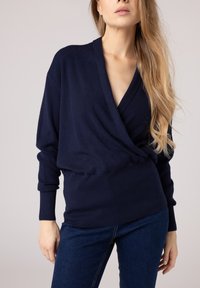 Granatowy sweter zapinany w miękkim, dzianinowym materiale z głębokim dekoltem w kształcie V, długimi rękawami i ściągaczami. Połączony z ciemnoniebieskimi dżinsami.