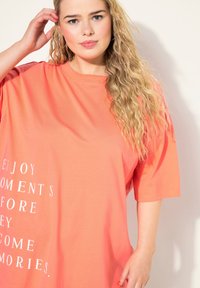 Camiseta de gran tamaño de color coral hecha de tela suave, con texto blanco que dice "DISFRUTA DE LOS MOMENTOS ANTES DE QUE SE CONVIERTAN EN RECUERDOS."