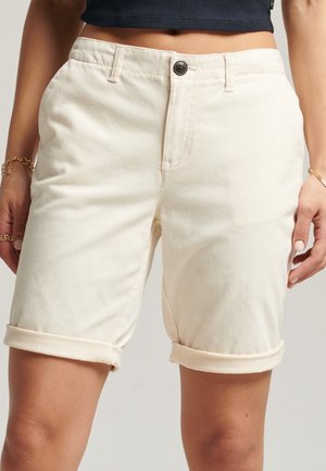 Mujer vestida con shorts de crema de tiro alto y dobladillo enrollado, con botón en la parte frontal y bolsillos, combinados con un top negro recortado y pulseras doradas.