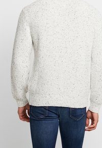 Weißer Strickpullover mit strukturiertem Muster, verziert mit schwarzen Sprenkeln. Lange Ärmel und ein gerader Saum, kombiniert mit dunkelblauen Jeans.