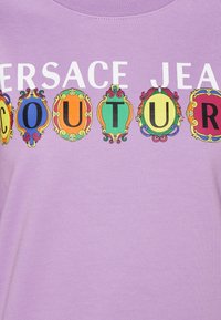 T-shirt lilas avec un grand texte "Versace Jeans Couture", arborant des motifs de cadre ornementaux colorés entourant certaines lettres. Tissu doux et lisse.