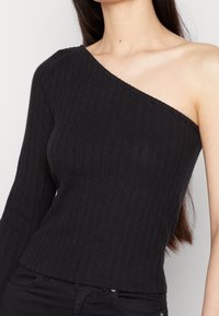 Top negro de punto acanalado con un hombro, diseño ajustado y un escote más amplio en el lado desocupado. Textura suave y material elástico.