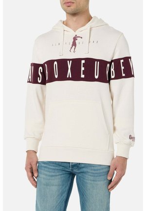 Mann trägt einen cremefarbenen Hoodie mit einem weinroten Streifen und dem Schriftzug "BOXEUR" über der Brust, kombiniert mit blauen Jeans, steht mit entspannten Händen seitlich am Körper.