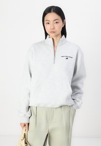 Pull en polaire gris avec un col zippé au quart, équipé d'une poche avant et d'un logo "New Balance" brodé en noir. Texture lisse.