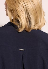 Granatowa koszula z klasycznym kołnierzykiem, z zaszewką z tyłu i małą metalową metką. Materiał wydaje się gładki i teksturowany.