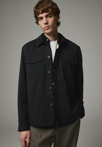 Sort jakke med knaplukning foran, to forlommer, krave og lange ærmer. Båret over en lysefarvet skjorte og beige bukser.