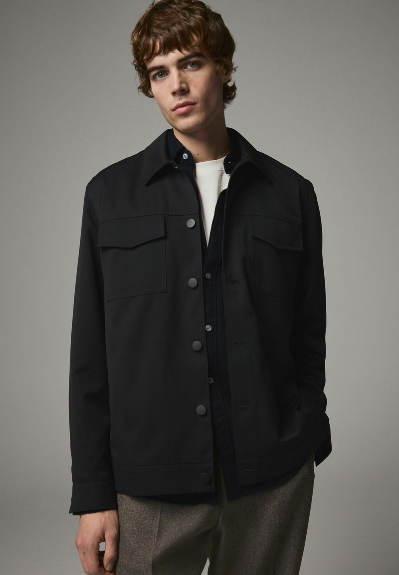 Sort jakke med knaplukning foran, to forlommer, krave og lange ærmer. Båret over en lysefarvet skjorte og beige bukser.