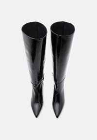 Botas negras hasta la rodilla de material brillante, con punta afilada y una silueta esbelta. Detalles decorativos cerca de la parte superior.