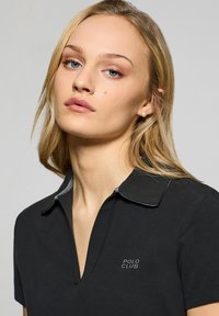 Svart polotröja med V-hals och korta ärmar, med en kontrasterande krage. Har "POLO CLUB" broderat på bröstet. Slät textur.