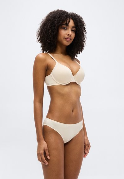 Γυναίκα με σγουρά μαλλιά που φοράει μπεζ σουτιέν και αντίστοιχο εσώρουχο, στέκεται μπροστά σε απλό φωτεινό φόντο.