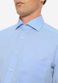 Camicetta azzurro chiaro realizzata in tessuto morbido; presenta un colletto strutturato, chiusura con bottoni e una tasca sul lato sinistro.
