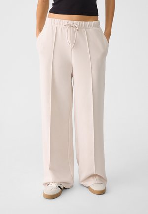 Osoba ubrana w luźne, jasnobeżowe spodnie z sznurkiem w pasie, z przednimi zakładkami, oraz białe trampki z gumowymi podeszwami, z rękami w kieszeniach.