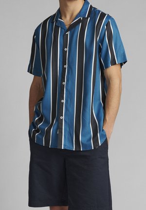 Mann trägt ein kurzärmeliges Hemd mit vertikalen blauen, schwarzen und weißen Streifen und dunkle marineblaue Shorts, Hände in den Taschen.