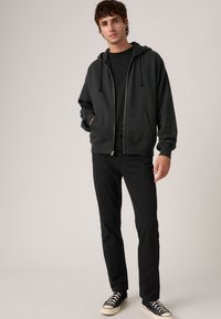 Μαύρο φούτερ με φερμουάρ και πλαϊνές τσέπες, φορεμένο πάνω από μαύρο πουκάμισο και συνδυασμένο με μαύρο παντελόνι και μαύρα αθλητικά παπούτσια, με άνετη εφαρμογή και υφή στο ύφασμα.