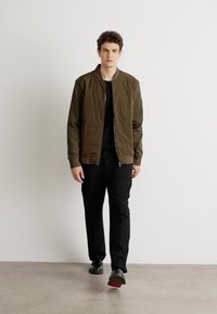 Olive green bomber jacket with a quilted texture, black t-shirt, black pants, and black shoes with red soles. Model walking against a neutral background.