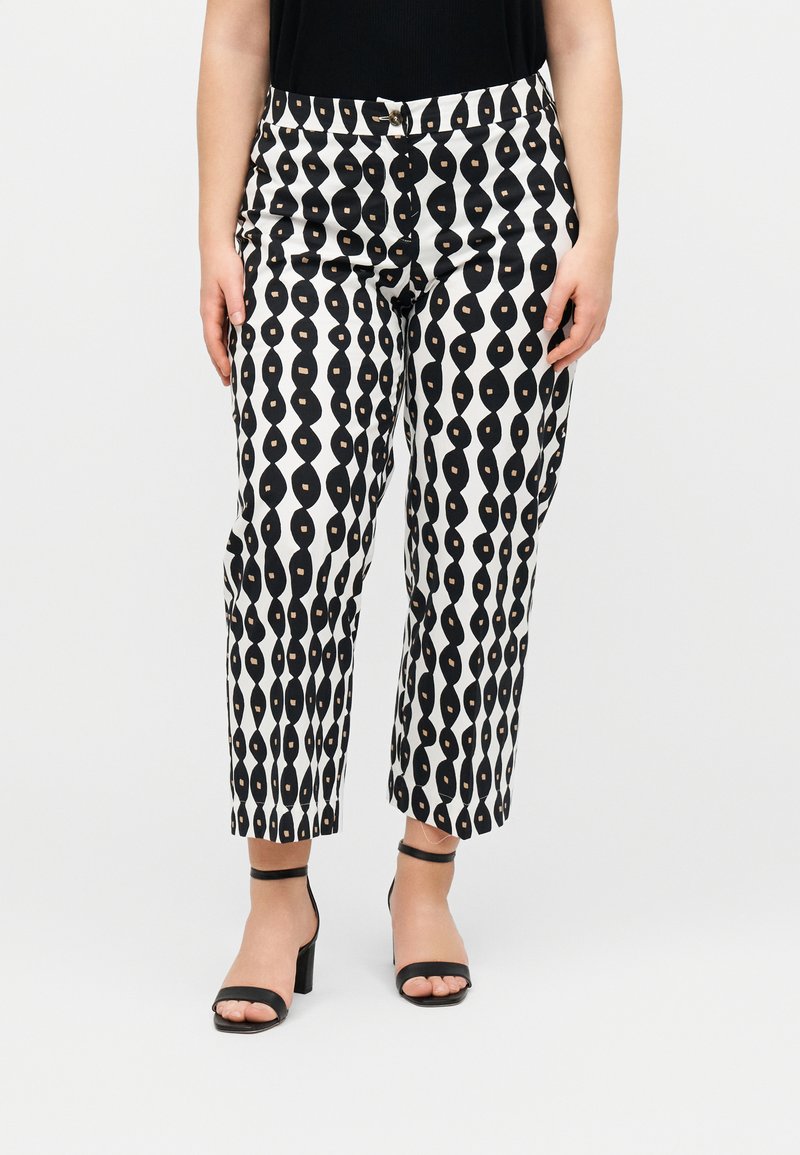 Γυναίκα που φοράει μαύρο και άσπρο παντελόνι cropped με μοτίβο και μαύρα σανδάλια με τακούνι μπροστά σε απλό φόντο.