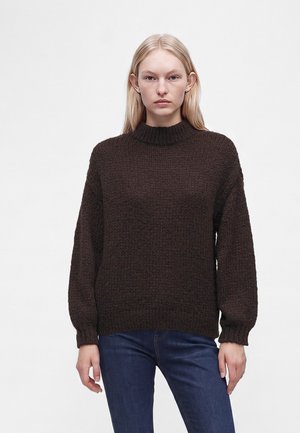 Femme blonde portant un pull en grosse maille marron foncé et un jean bleu foncé sur un fond clair uni.