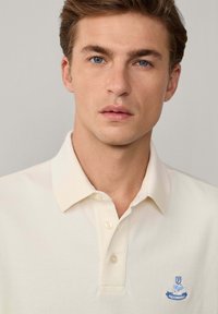 Polo shirt di colore crema chiaro con una texture morbida, dotato di colletto e tre bottoni. Un piccolo logo blu ricamato si trova sul lato sinistro del petto.