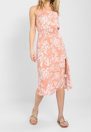 Mujer vistiendo un vestido midi color durazno con estampado floral blanco, cintura fruncida, abertura lateral y sandalias planas doradas con tiras, de pie contra un fondo blanco.