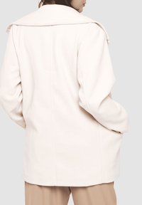 Abrigo oversized color beige claro con un cuello ancho, textura de tela suave y un diseño estructurado. Presenta un corte recto y un minimalismo en los detalles.