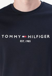Camiseta de algodón azul marino con texto bordado en blanco "TOMMY HILFIGER" y "EST. 1985" junto con un acento de logotipo rectangular en rojo, blanco y azul.