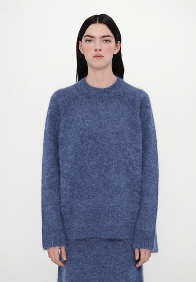 Pull en laine bleu avec un col rond, des manches longues et une texture douce et pelucheuse. L'ourlet est légèrement plus long à l'arrière.