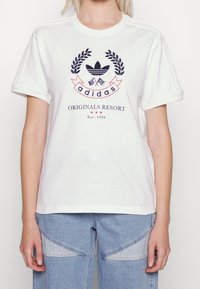 Witte katoenen T-shirt met een marineblauw en rood gedrukt logo met lauweraccenten en de tekst "adidas ORIGINALS RESORT Est. 1949."