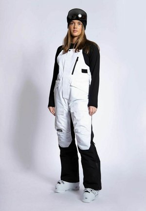 Ung kvinde iført sort og hvid snowboardtøj, sort hue, goggles og hvide snowboardstøvler, stående mod en ensfarvet baggrund.
