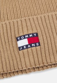Gorro de punto beige con textura acanalada y una pequeña etiqueta azul marino con "TOMMY JEANS" y un logo en rojo, blanco y azul.