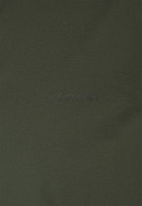 Tejido verde oliva con una textura suave, con el logotipo "Calvin Klein" sutilmente colocado en negro. El diseño es minimalista y monocromático.